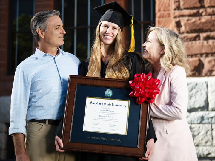 blonde haired girl in a black grad cap with yellow tassel and black gown holding a diploma frame with her father smiling at her on her left and her mom on her right smiling at her.