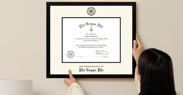 black haired woman hanging her Phi Kappa Phi certificate frame with a black moulding on a white wall.