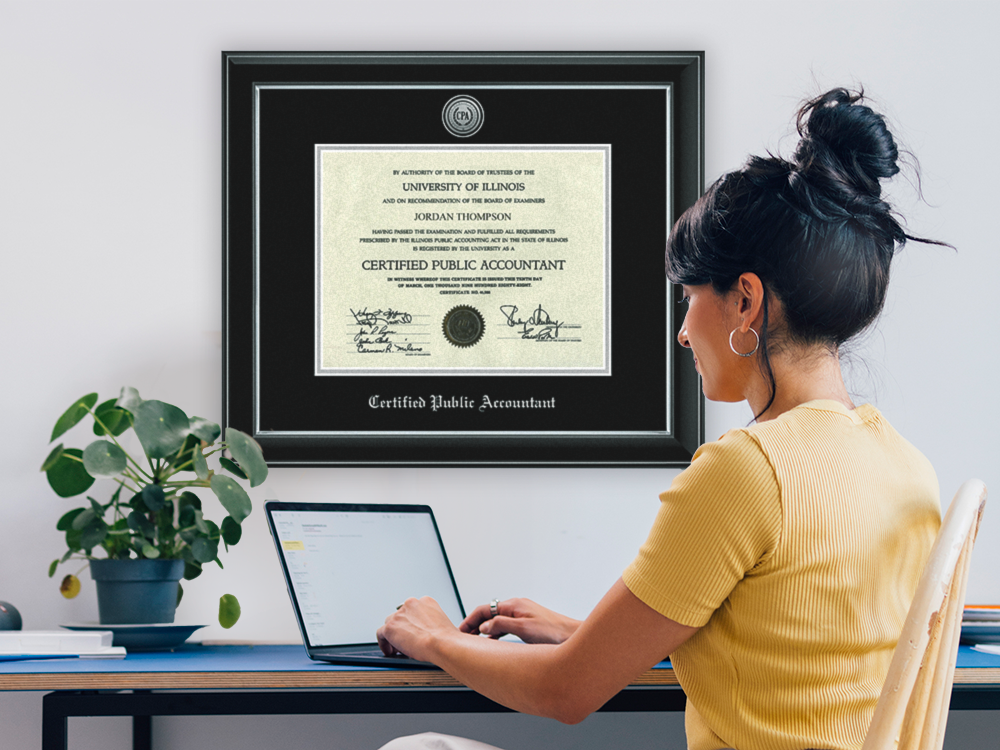 Professional certificate frame on a wall beside a woman working on a computer