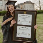 Smiling woman in graduation attire on Kansas State University campus holding a KSU double diploma frame.