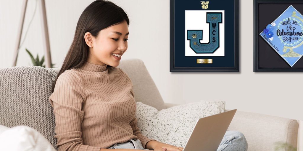 smiling teen typing on laptop with varsity letter frame and grad cap frame on wall