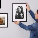 Woman hanging black photo frames of herself and fiance on gray wall