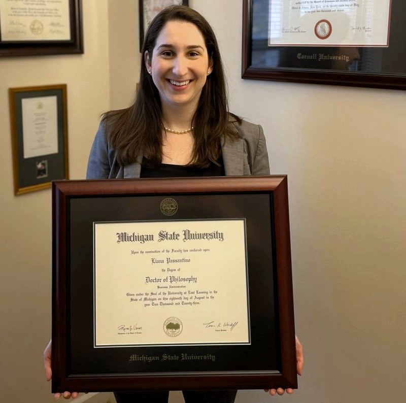 smiling woman wearing pearls holding michigan state diploma frame in office