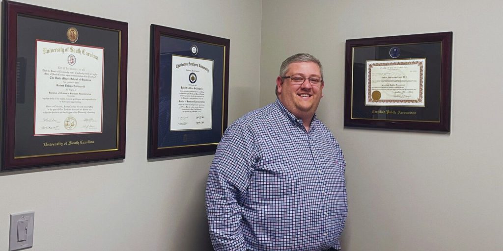 man standing next to his charleston southern and USC diploma frames and CPA certificate frame on gray wall