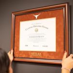 back of woman's head who is hanging UT Austin diploma frame on wall