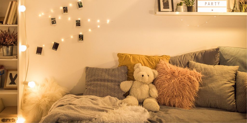 cozy dorm room with pink throw pillow and teddy bear on bed with fairy lights in background