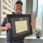 A smiling man holding his state of washington law degree frame in front of office window