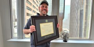 A smiling man holding his state of washington law degree frame in front of office window