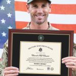smiling soldier holding army certificate frame in front of american flag