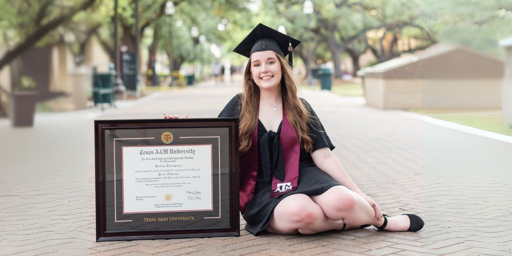 Texas A&M graduate wearing regalia posing with a custom Texas A&M diploma frame