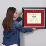 woman janging a custom diploma frame on wall next to plant and custom photo frame