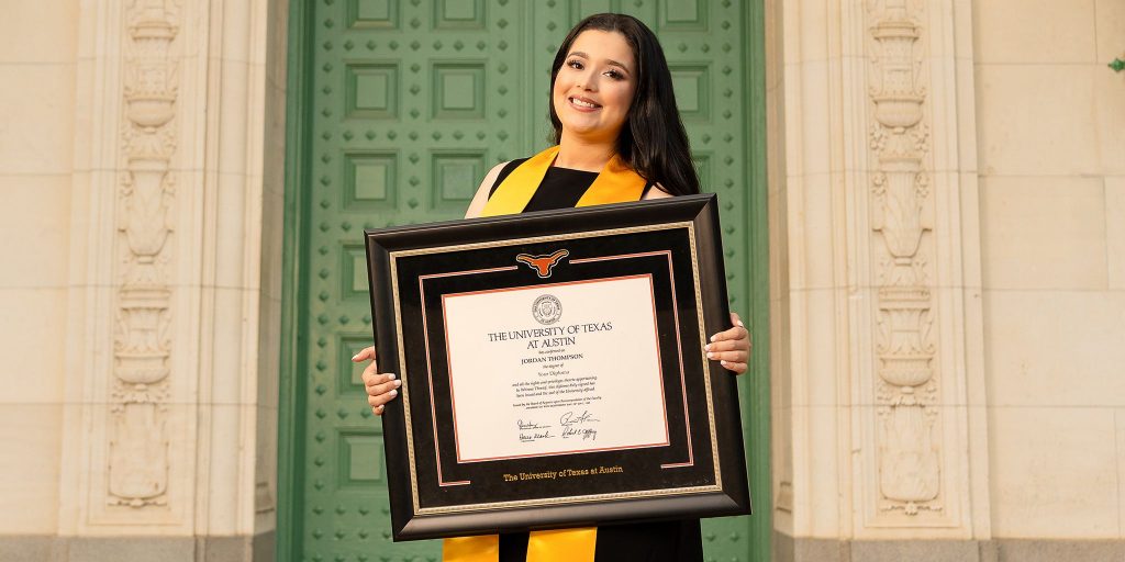 Smiling graduate in regalia holding a custom UT Austin diploma frame