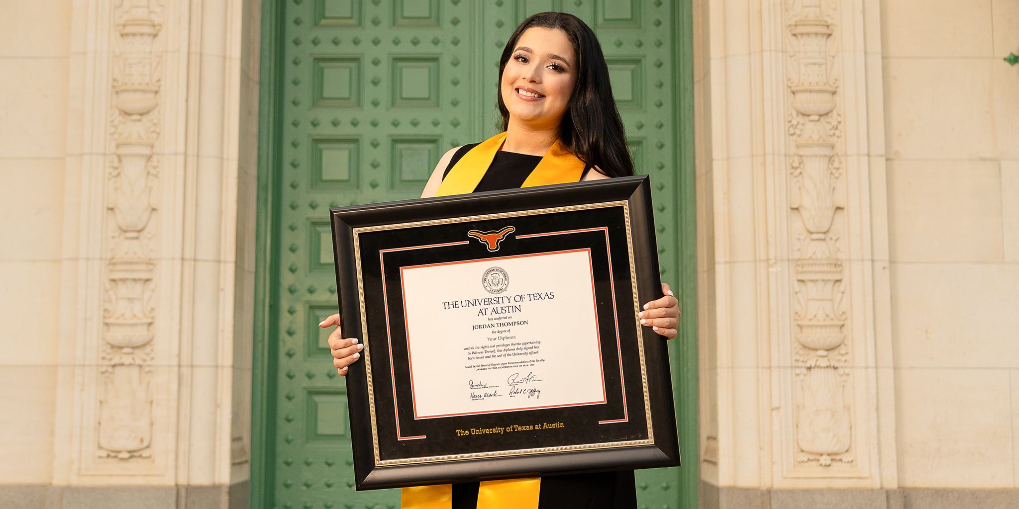 Smiling graduate in regalia holding a custom UT Austin diploma frame