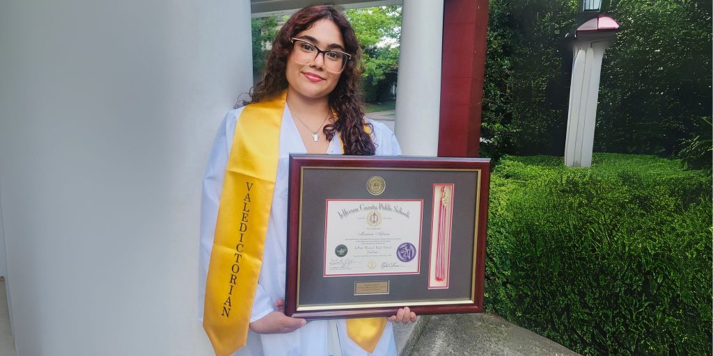 Graduate wearing gold valedictorian stole and holding tassel diploma frame in front of column outdoors.