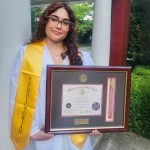 Graduate wearing gold valedictorian stole and holding tassel diploma frame in front of column outdoors.