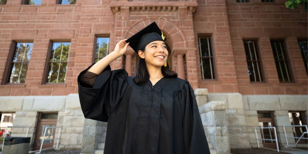 smiling graduate in black gown and cap in front of college building