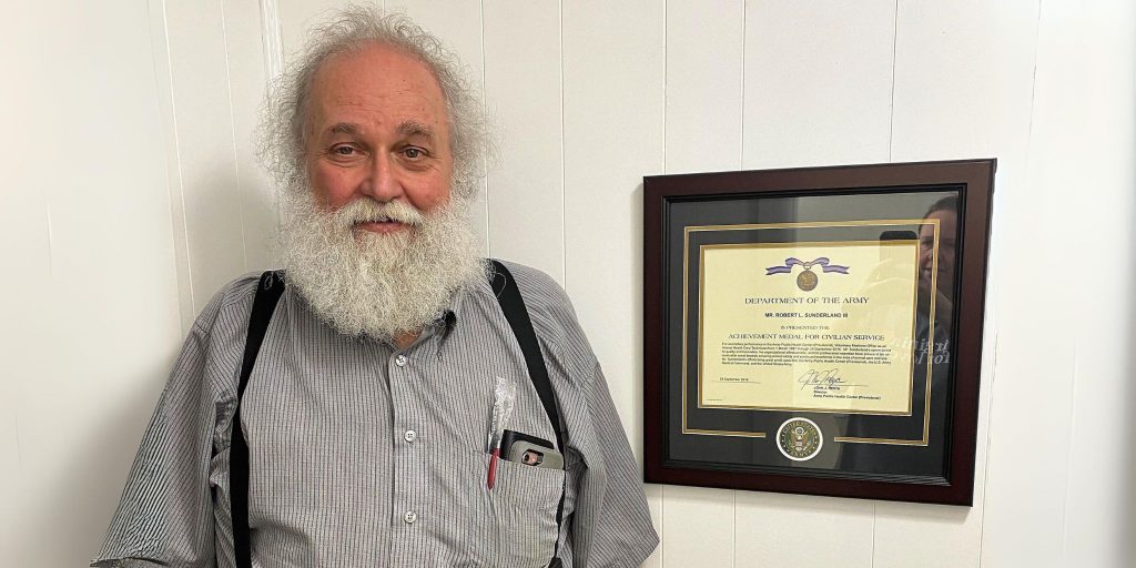 Grandpa with white beard standing next to Army certificate frame on wall