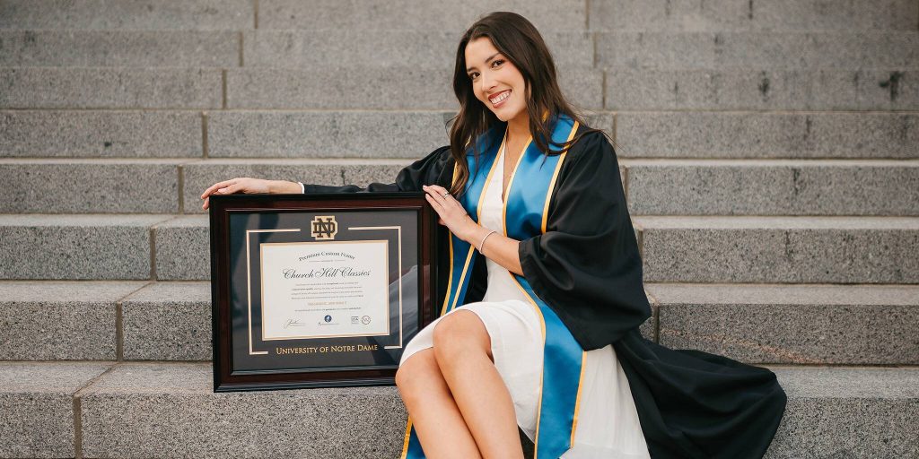 smiling notre dame graduate sitting on stairs posing with custom Notre Dame diploma frame