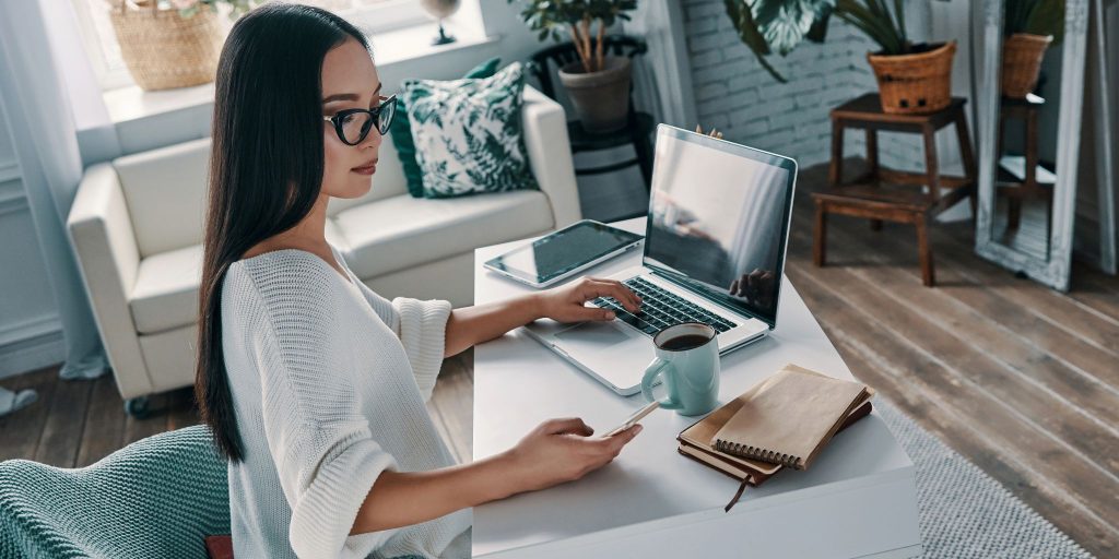 woman working at desk in living looking at phone and typing on laptop