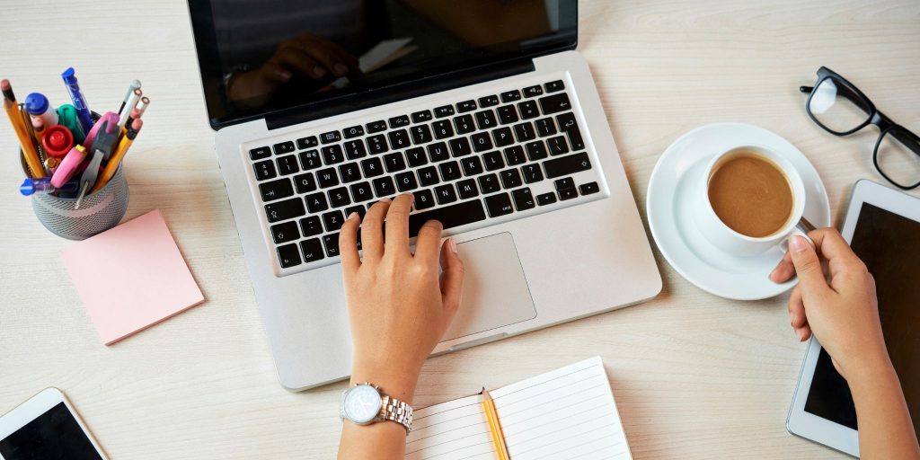 closeup of desk with one hand working on laptop and the other holding cup of coffee with pencil holder, sticky notes, and glasses on top of desk