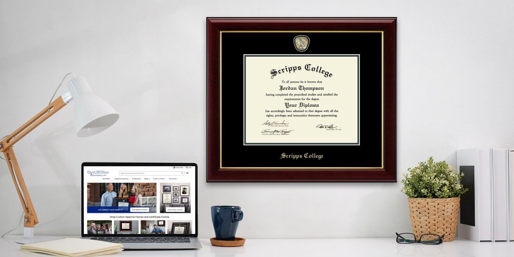 Scripps College diploma frame hung on a wall behind a desk with a lamp, laptop, and plant