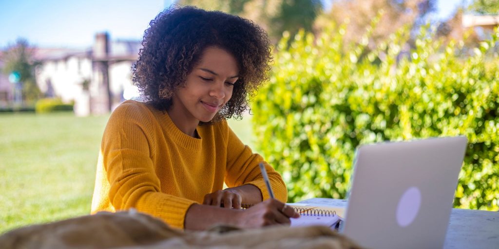 college student outdoors at table writing in notebook in front of laptop