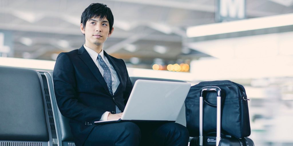 man in suit on laptop sitting at airport next to travel bag and suitcase