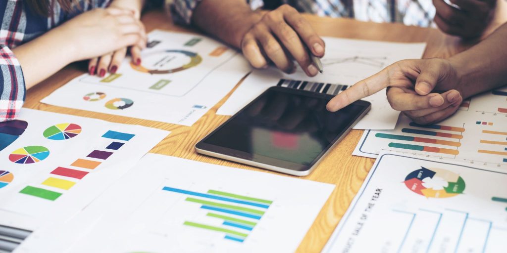 Closeup of table with printouts of bar charts and circle graphs and hands pointing to an iPad.