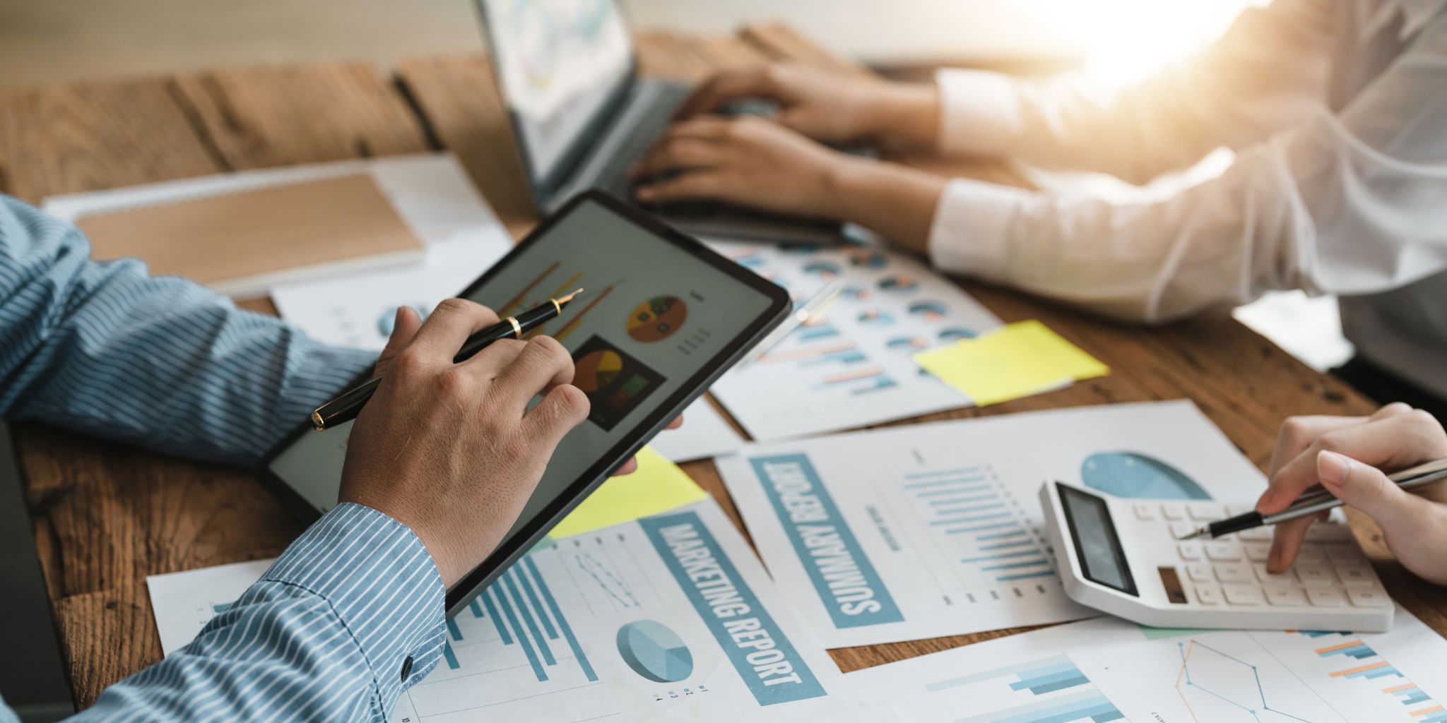 Closeup of hands on an iPad with pen over a desk covered with marketing reports and bar graphs.