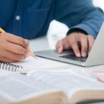 closeup of person holding pencil and writing in notebook on desk with other hand on a laptop and an open book in the foreground.