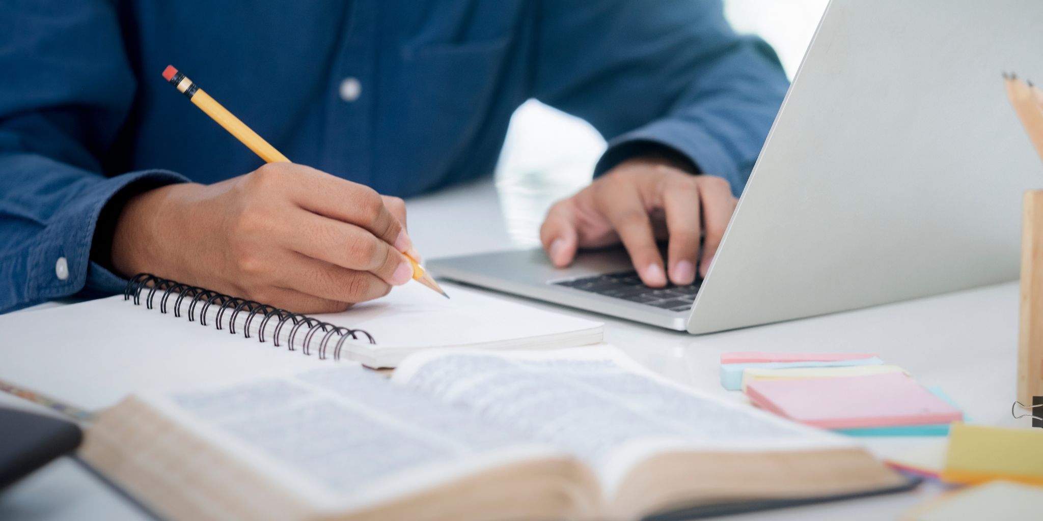 closeup of person holding pencil and writing in notebook on desk with other hand on a laptop and an open book in the foreground.