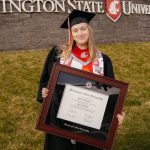 Graduate on campus in full regalia holding her Washington State University diploma frame.
