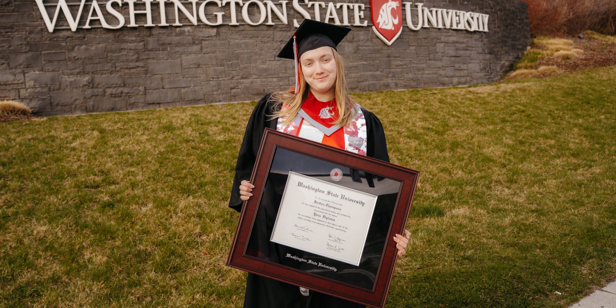 Graduate on campus in full regalia holding her Washington State University diploma frame.