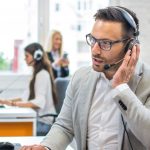 Man in gray suit in busy office wearing headset in front of laptop and talking into headset microphone.