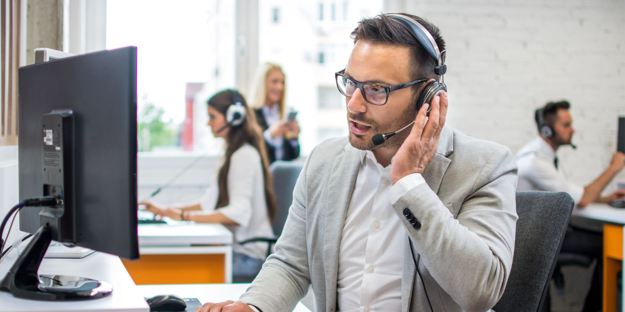 Man in gray suit in busy office wearing headset in front of laptop and talking into headset microphone.