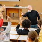 Professor in lecture hall standing in front of college students raising their hands to answer his question.