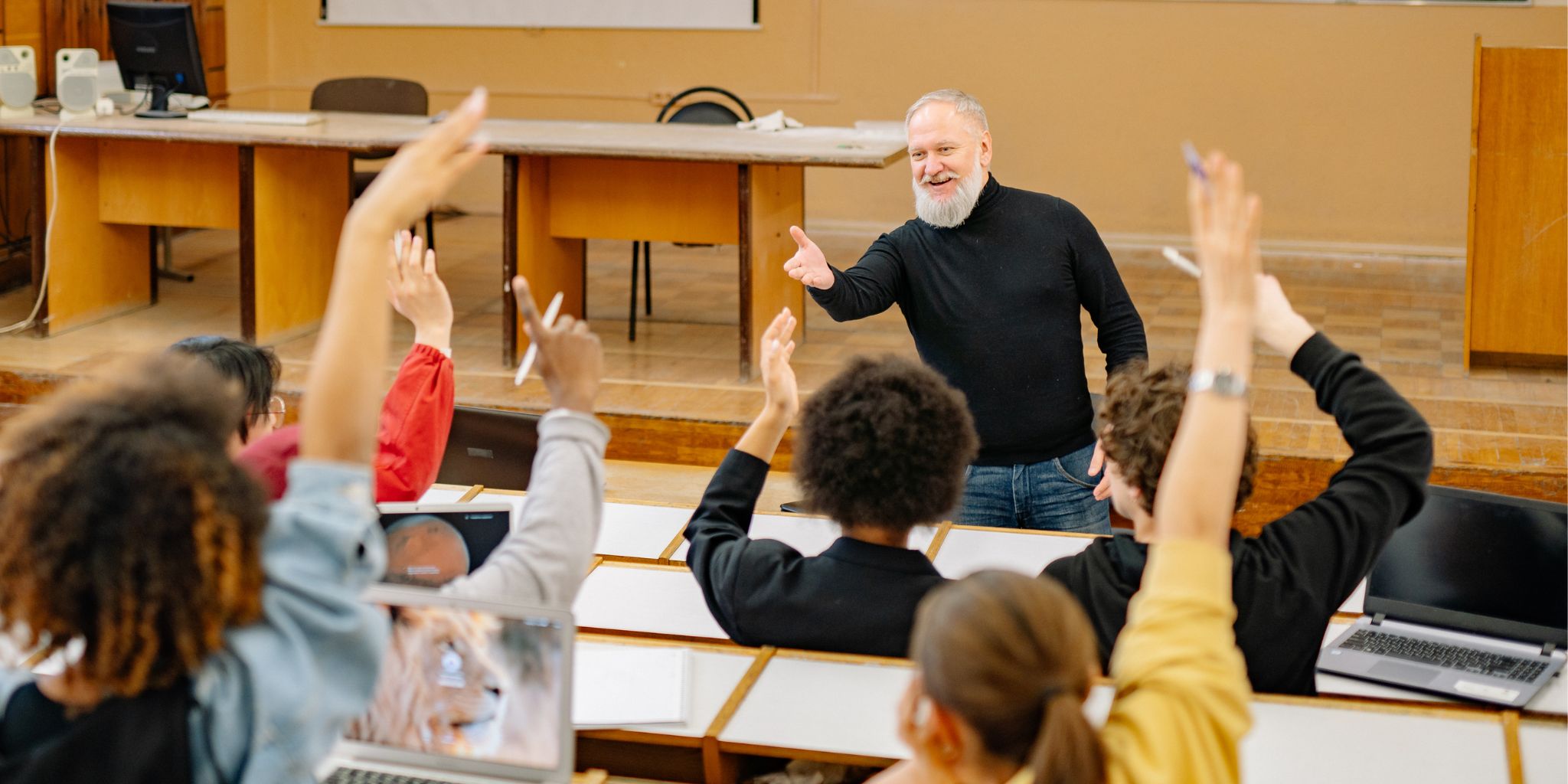 Professor in lecture hall standing in front of college students raising their hands to answer his question.
