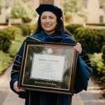 Graduate in full regalia holding Notre Dame diploma frame on campus.