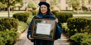 Graduate in full regalia holding Notre Dame diploma frame on campus.