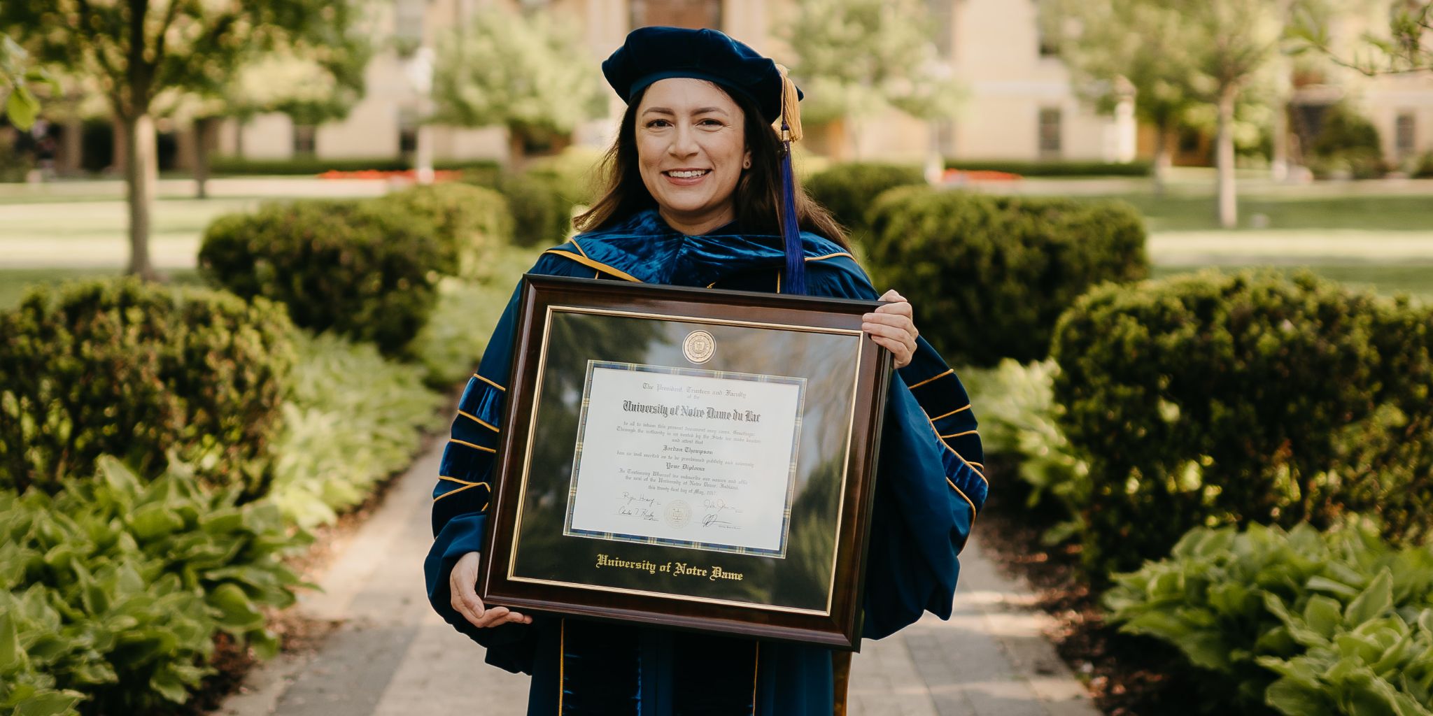 Graduate in full regalia holding Notre Dame diploma frame on campus.