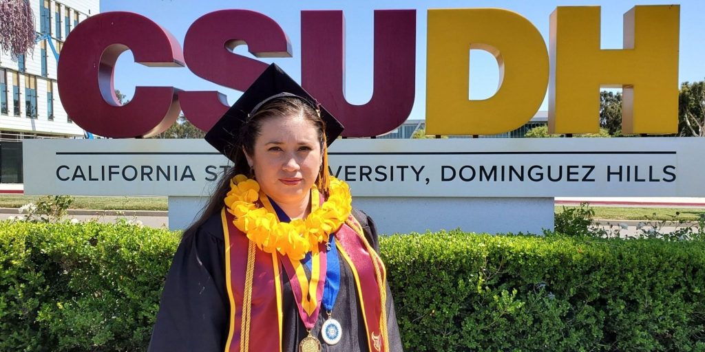 Graduate in full regalia with graduation stole, medallions, and lei standing in front of CSUDH sign.