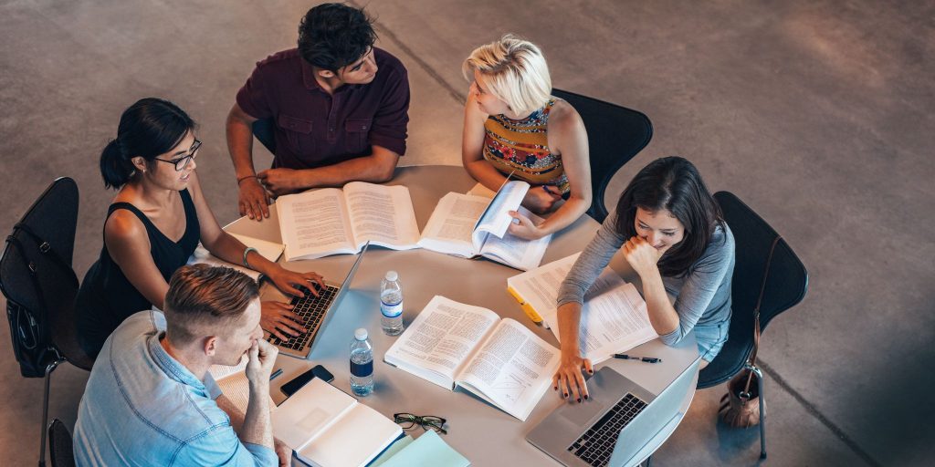 Five college students studying together at the same table with books, laptops and water bottles.