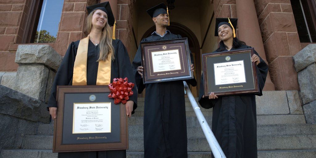 Three grads on university steps holding their college diploma frames.