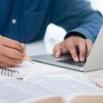 Closeup of person in blue shirt with pencil in hand writing in notebook as they work on laptop at desk.
