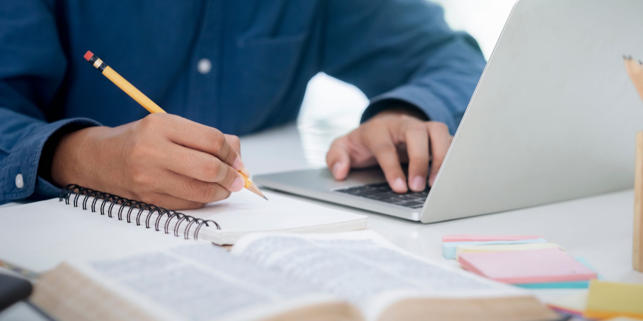 Closeup of person in blue shirt with pencil in hand writing in notebook as they work on laptop at desk.