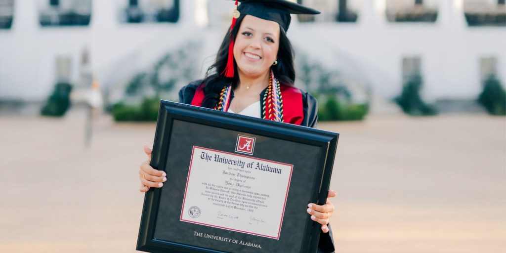 Smiling university of alabama graduate wearing regalia and holding a custom diploma frame