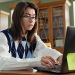 Student in glasses and argyle sweater working at computer in college library.