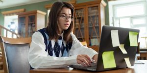 Student in glasses and argyle sweater working at computer in college library.