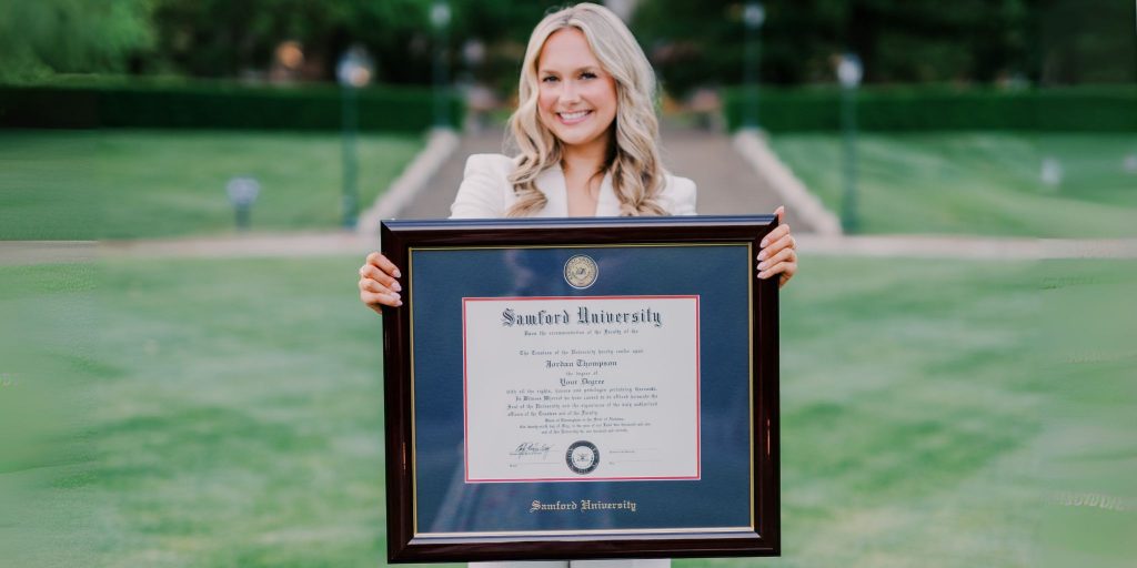Smiling woman on campus holding a large Samford University diploma frame.