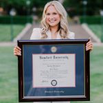 Smiling woman on campus holding a large Samford University diploma frame.